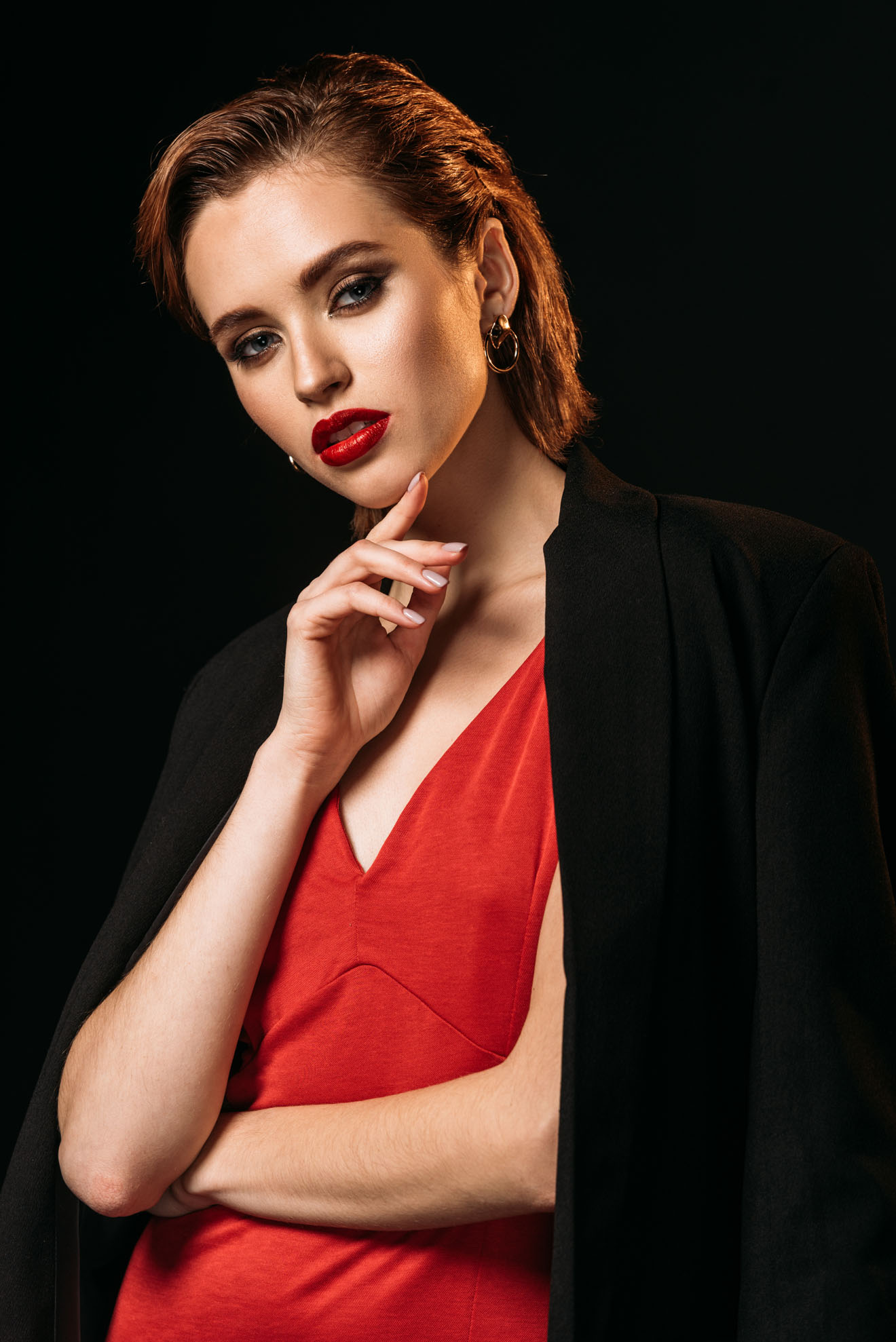 attractive girl in red dress and black jacket touching chin and looking at camera isolated on black